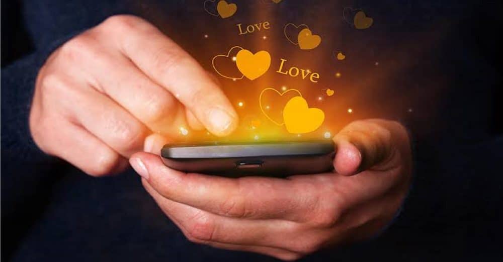 An anonymous person in a dimly lit room holding a smartphone that displays glowing love icons on a dark background.